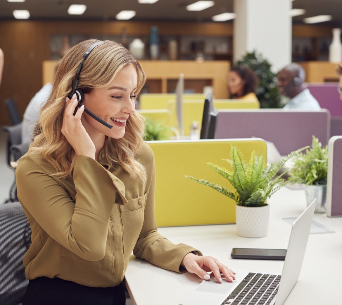Businesswoman Wearing Phone Headset Talking To Caller In Busy Customer Services Centre